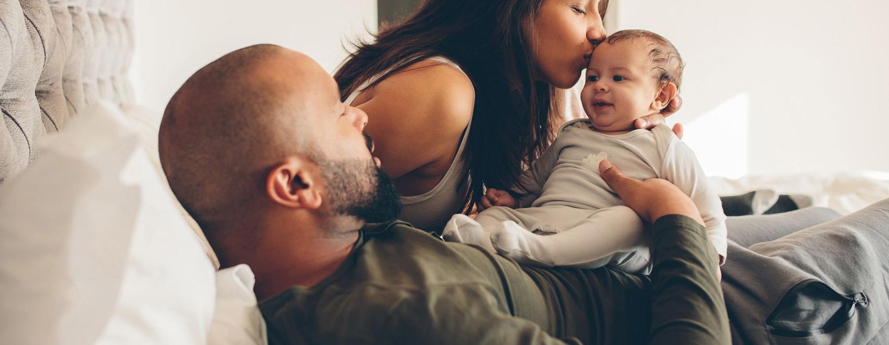 mother kisses her infant while father lies in bed and holds him